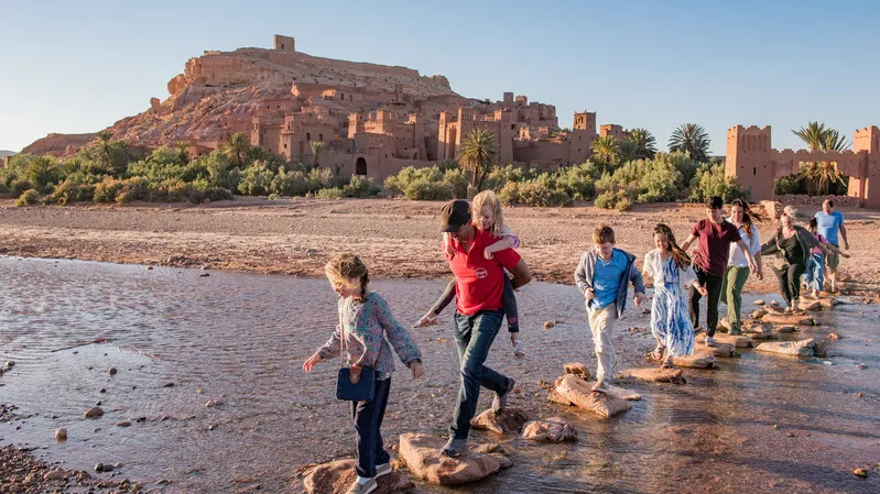 A group of tourists crossing a river in front of a kasbah while their Family Tours in Morocco