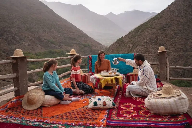 A group of touristssitting on top of a blanket in the middle of the Atlas Mountains with local guide drinkink Moroccan tea while their family tours in Moroccoo