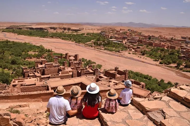 A group of tourist sitting on top of a stone wall watching old villages and kasbah and palm trees while their family tours in Morocco 