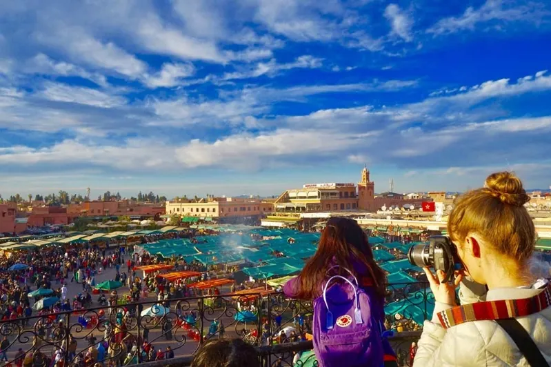 Tourists capturing moments at the vibrant Marrakesh market, surrounded by colorful stalls and lively atmosphere.While Moroccan Travel Agency