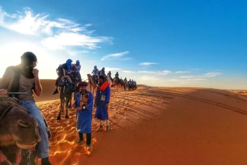 Several people mounted on camels traverse the expansive desert, surrounded by golden sand dunes and bright sunlight.While Morocco desert tour