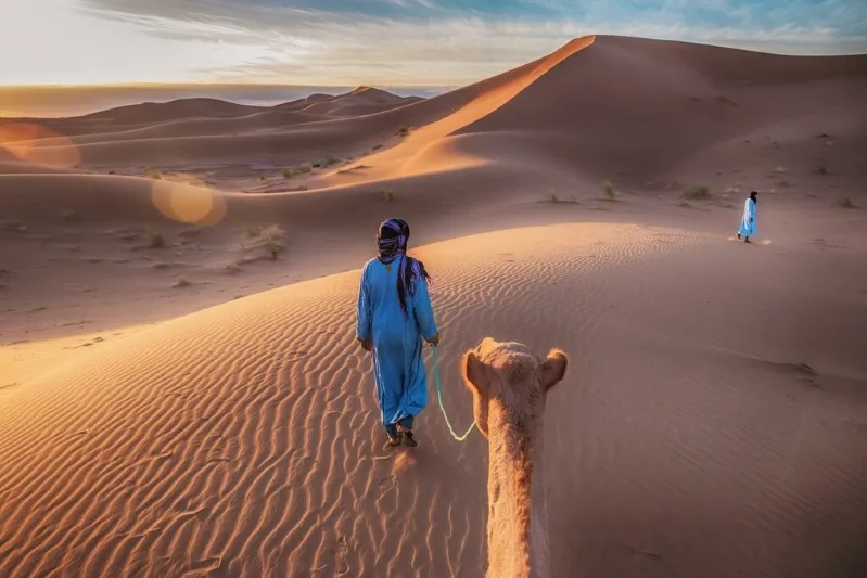  A man and a camel traverse the desert landscape as the sun sets, casting warm hues across the sandy terrain. While luxury morocco desert tour 