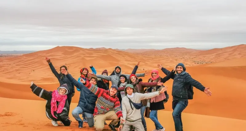  A cheerful group of people standing together in the desert, capturing a moment with smiles against a sandy backdrop.While Morocco desert tour