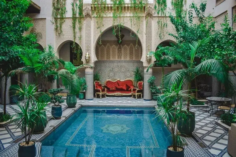 A hotel courtyard featuring a serene pool surrounded by greenery and a vibrant red couch for relaxation.While Morocco travel deals