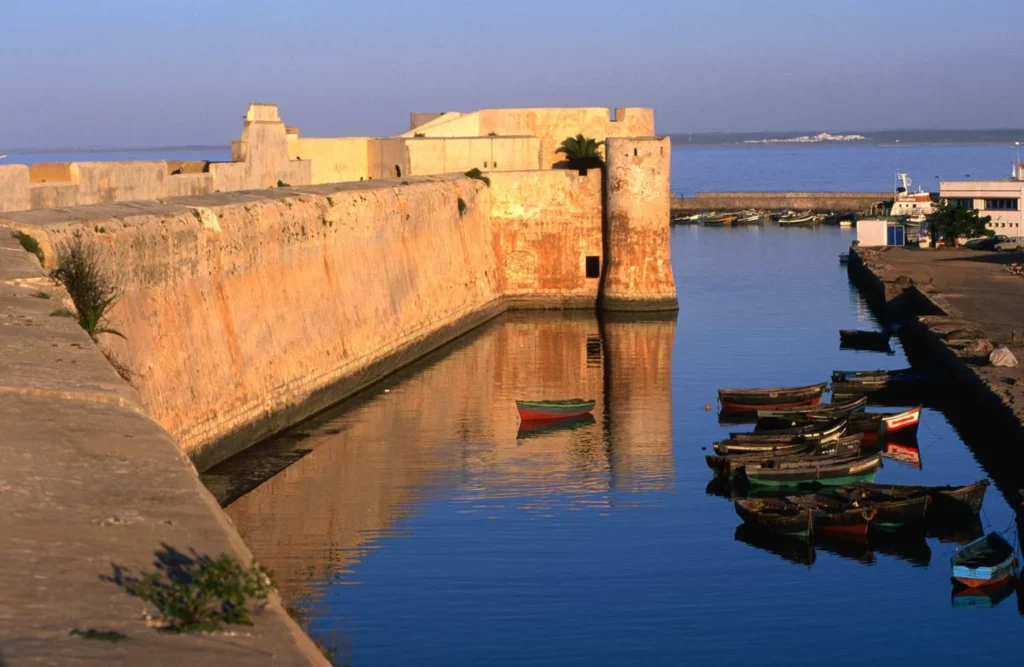 Boats are docked in the water beside a stone wall, creating a serene waterfront scene. A Day Trip to El Jadida