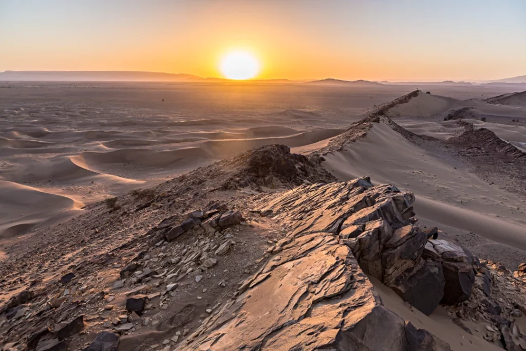 The sun sets over the Sahara Desert, casting warm hues across the vast, sandy landscape.