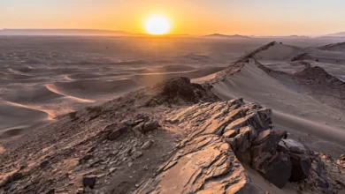 The sun sets over the Sahara Desert, casting warm hues across the vast, sandy landscape.