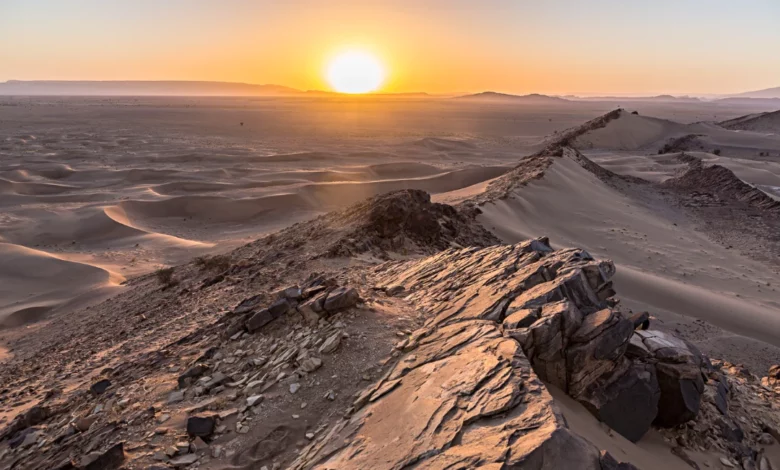 The sun sets over the Sahara Desert, casting warm hues across the vast, sandy landscape.
