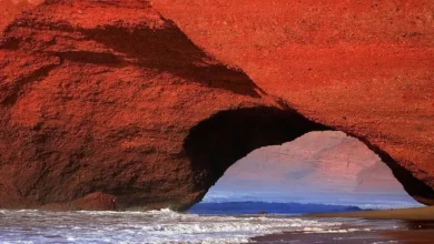 A red rock arch stands on the beach, with waves crashing against the shore beneath a clear blue sky.