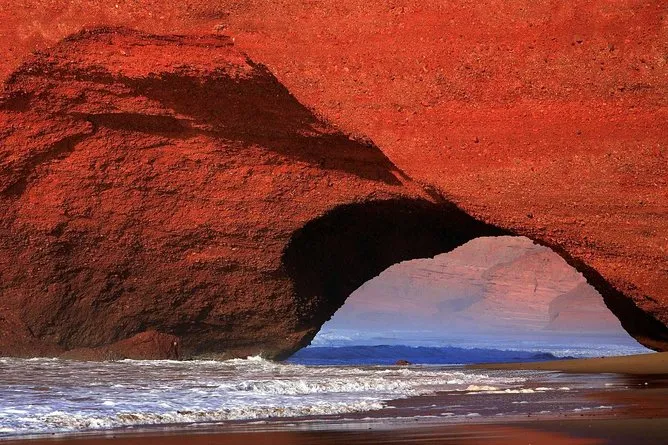 A red rock arch stands on the beach, with waves crashing against the shore beneath a clear blue sky.