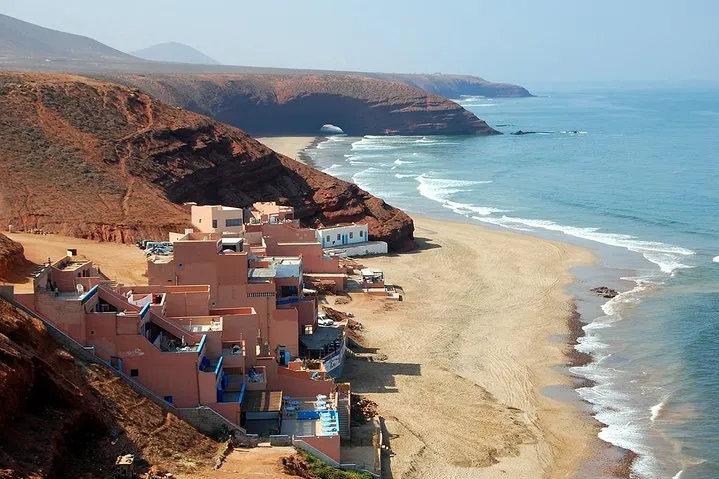 A scenic beach with houses perched on a cliffside, overlooking the ocean waves and sandy shore below. From Agadir, enjoy a Southern Coastline Exploration to Sidi Ifni & Mirleft Day Trip: a coastal adventure full of charm and ocean views.