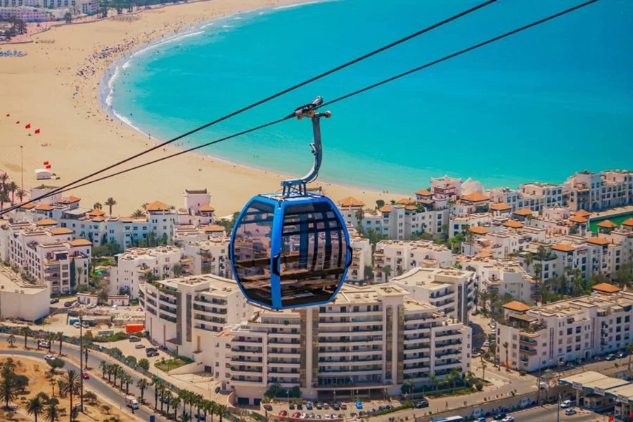 A cable car glides over a beach, offering a view of the ocean below.7 Days Agadir to Imperial Cities & Desert Tour