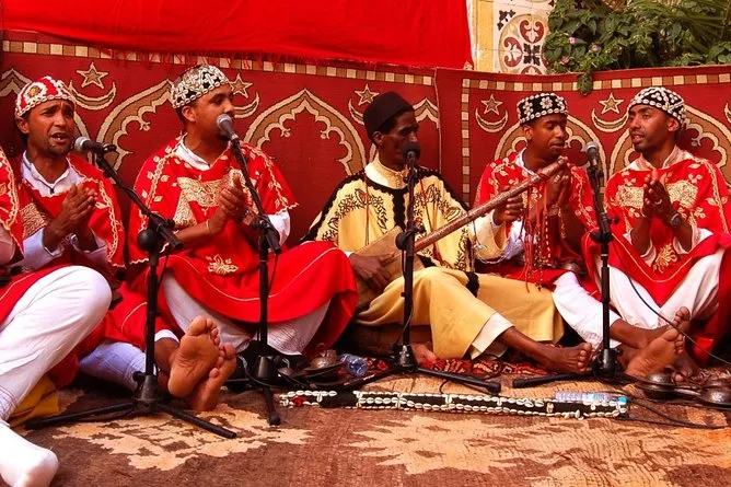 A group of men in traditional attire sits together around a microphone, engaged in conversation or performance.