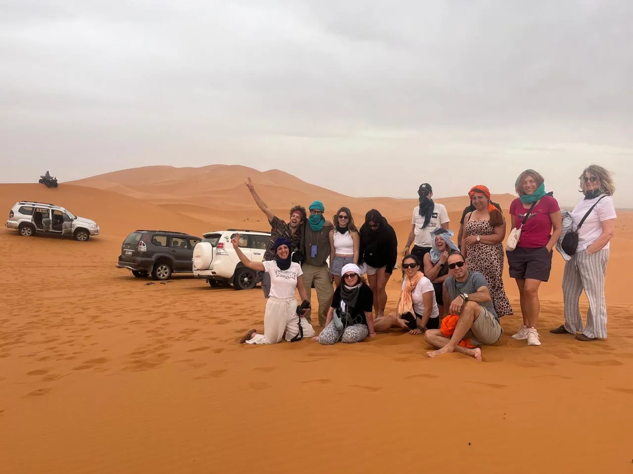 Group of people smiling and posing for a photo against a desert backdrop with sand dunes and clear blue sky. 4 Days Agadir to Desert & Ouarzazate