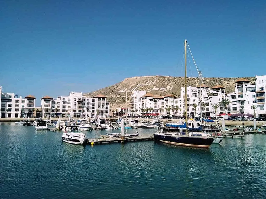 Boats are docked in a harbor with a mountain backdrop, creating a scenic coastal view. 4 Days Agadir to Desert & Ouarzazate
