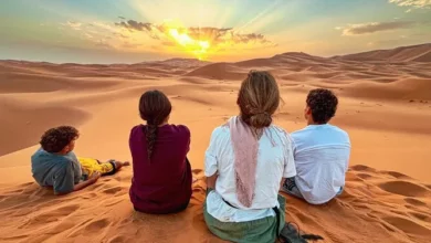 Three people sitting on the sand in a vast desert, surrounded by dunes under a clear blue sky.