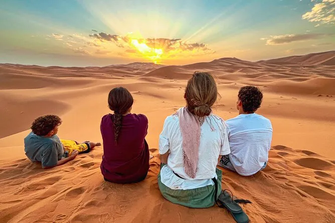 Three people sitting on the sand in a vast desert, surrounded by dunes under a clear blue sky. 