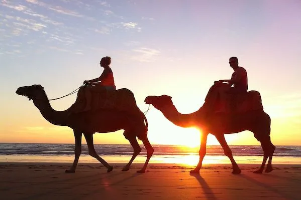 Two people ride camels along the beach at sunset, with vibrant colors reflecting on the water and sand.