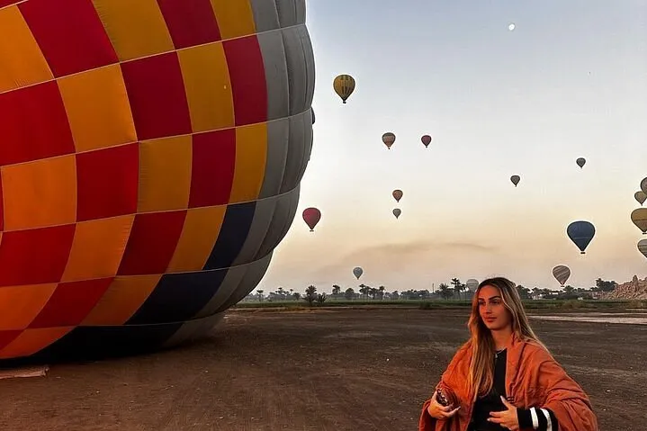 A woman stands smiling in front of a colorful hot air balloon against a clear blue sky.