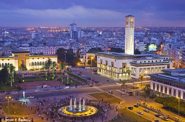 A cityscape featuring a prominent clock tower and a central fountain surrounded by buildings and greenery.
