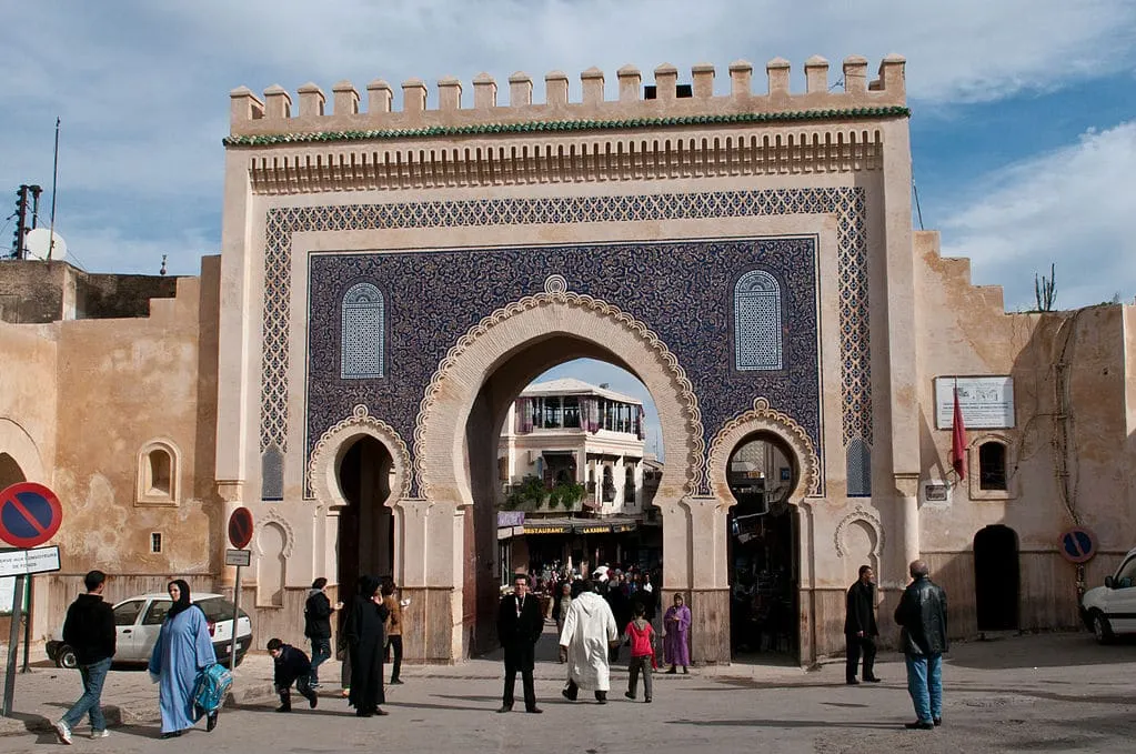 People walk through the entrance of a historic city gate, surrounded by stone walls and greenery.8 Days Tour from Fes