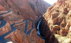 A winding mountain road alongside a river flowing through a lush valley, surrounded by towering peaks. 5 Days Fes to Marrakech via Desert & Dades Valley