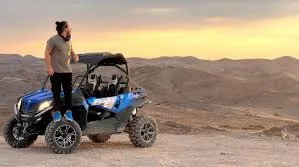 A man stands on a blue UTV in a vast desert landscape under a clear sky.