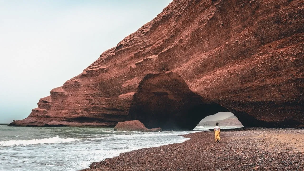 A person stands on the beach beside a large rock formation, with waves gently lapping at the shore in the background.