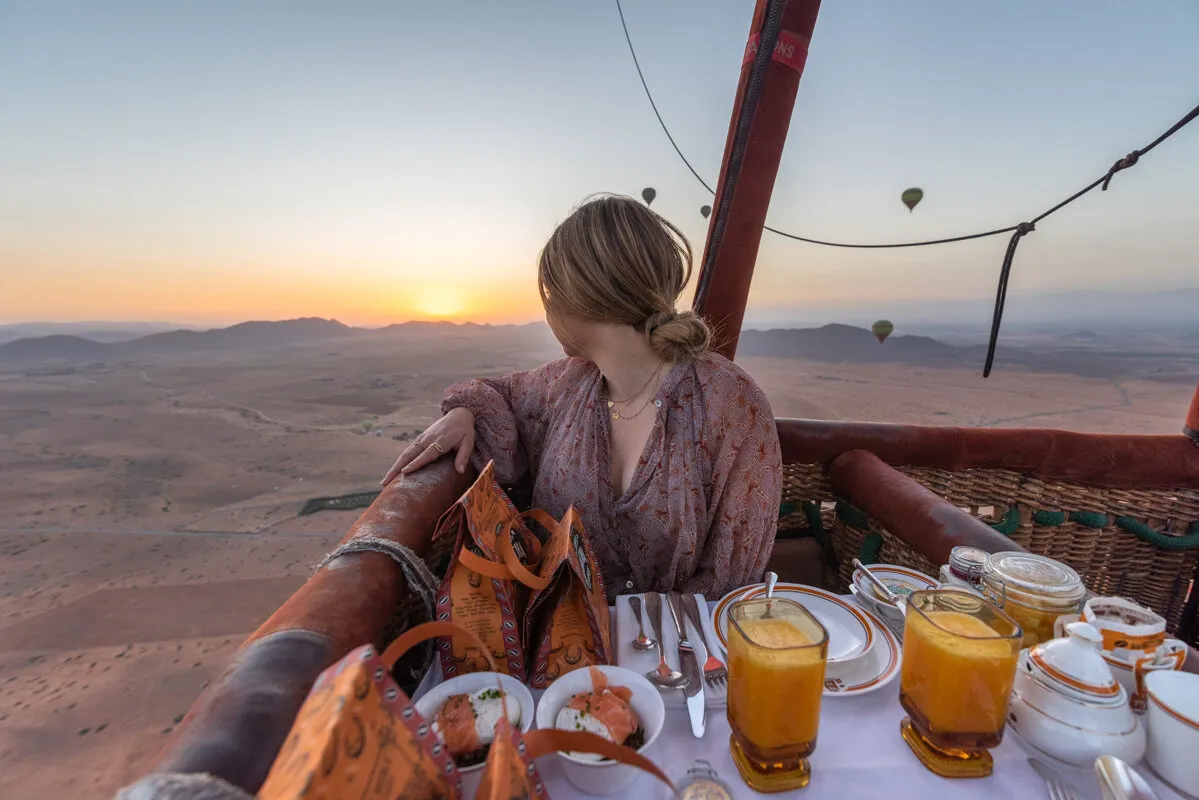 A woman sits at a table with food and drinks inside a hot air balloon, enjoying a unique dining experience in the sky.Vol en montgolfière au-dessus de Marrakech