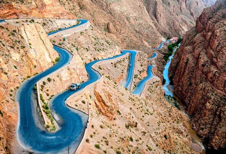 A winding mountain road alongside a vibrant blue river, surrounded by lush greenery and rocky terrain.
