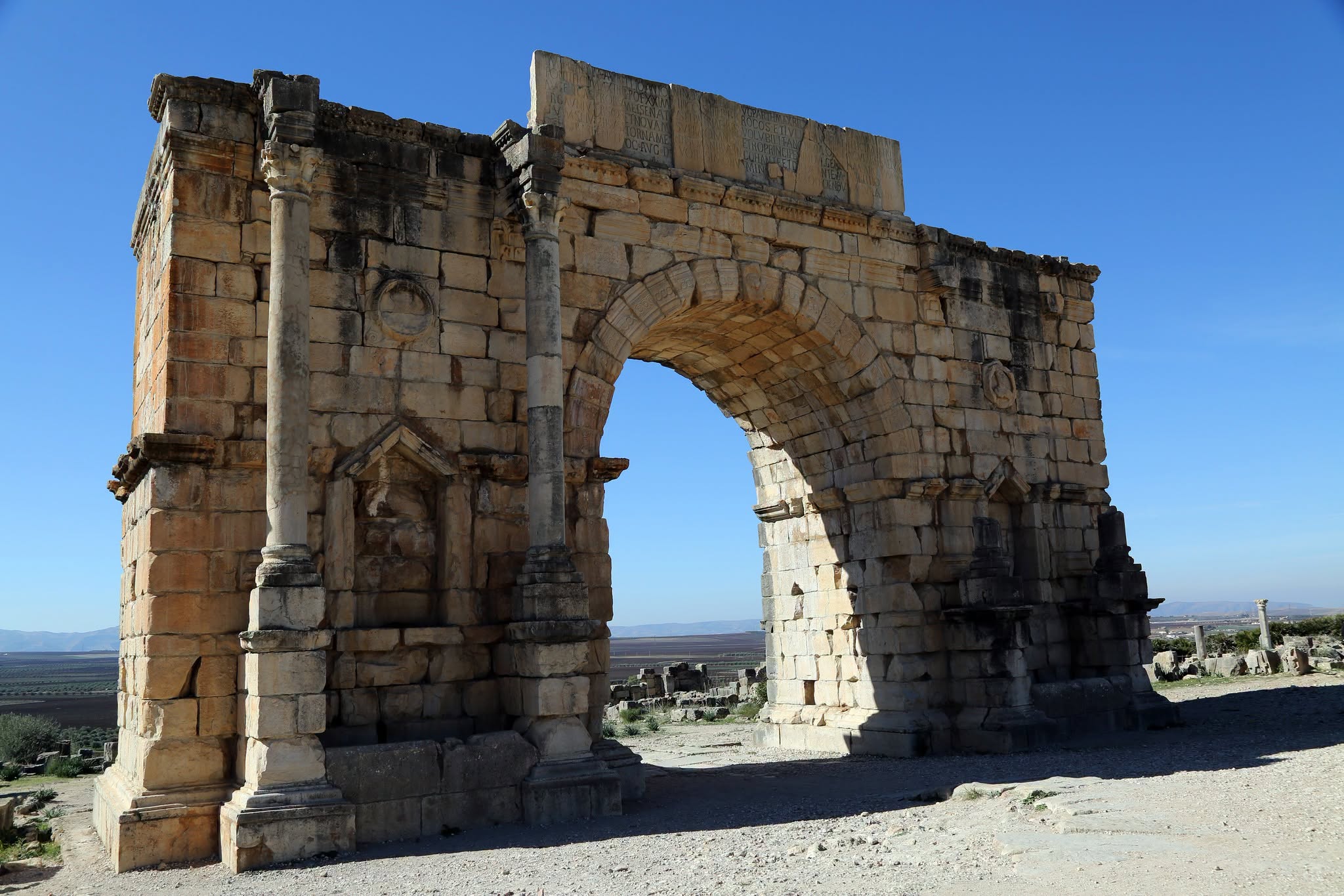 The Arch of Caracalla at Volubilis, seen from the path leading up to it, with open fields behind